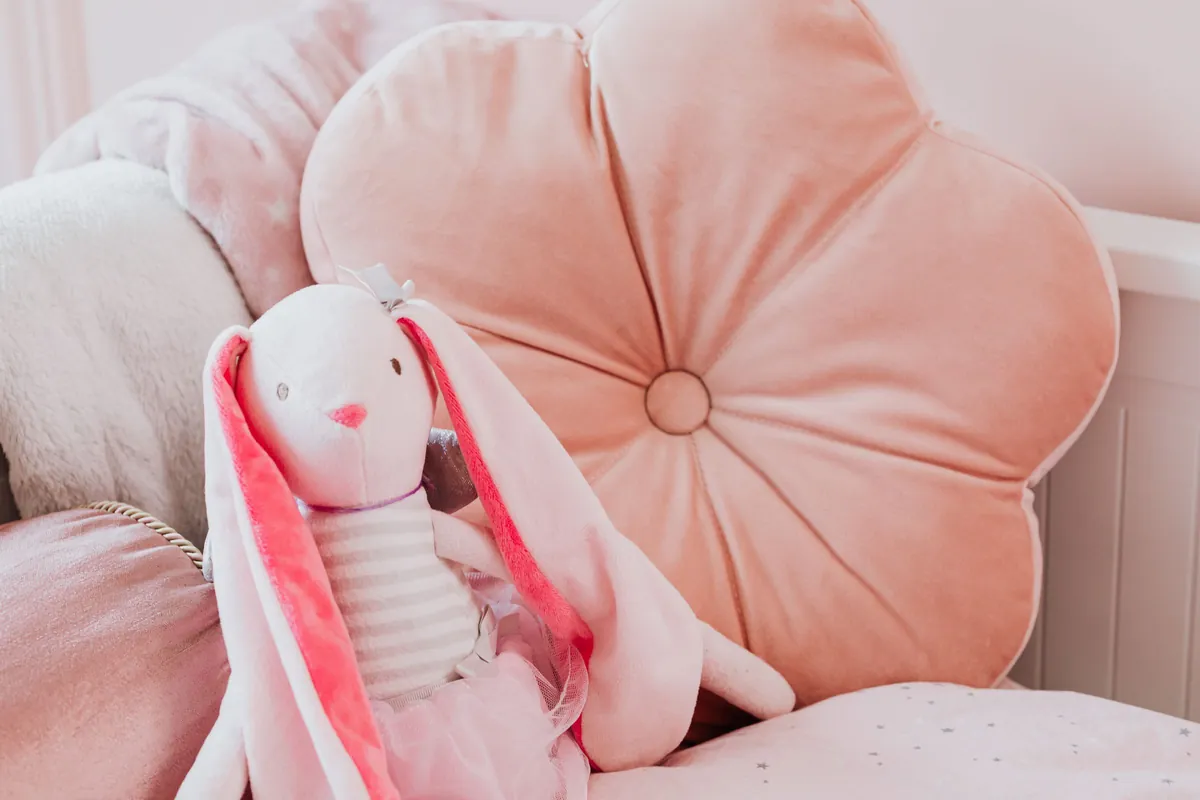 Oudroze bloemvormig kussen en een knuffelkonijn op een kinderbed tegen een zachtroze muur